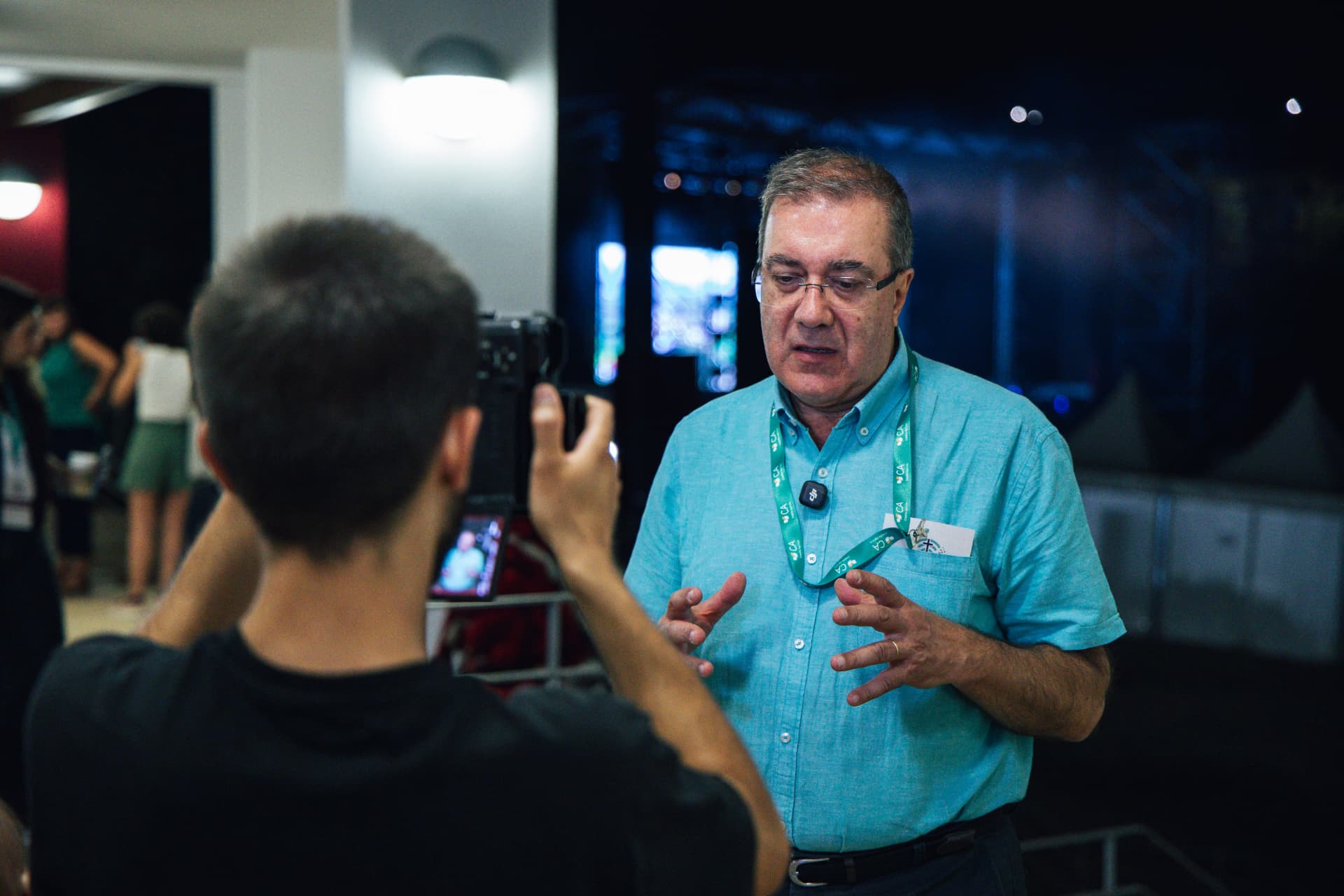 Sacerdote é entrevistado diante das câmaras no corredor envidraçado